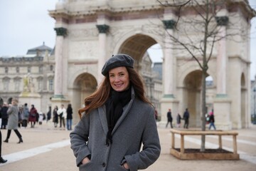 A cheerful woman wearing a gray coat and beret poses near the Arc de Triomphe du Carrousel in Paris, surrounded by historic architecture and a lively atmosphere.
