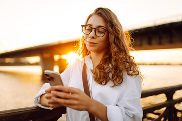 Fototapeta premium A young caucasian woman using her smartphone while walk on embankment. Concept of vocational, blogging, technology, weekend.