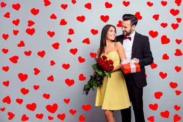 Portrait with copy space of attractive couple in formal wear, woman in dress holding bouquet of red roses, man in tux embracing her from back side and presenting a packaging red case with gift