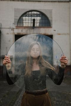Woman with eyes closed holding round glass pane and standing near building