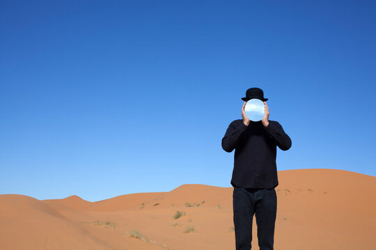 Morocco, Merzouga, Erg Chebbi, man wearing a bowler hat holding mirror in front of his face in desert dune