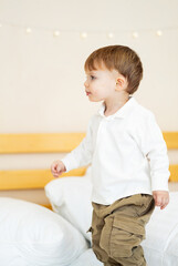 Naklejka premium Toddler playing and exploring indoors. A young child joyfully stands on a bed, looking around with curiosity in a bright room filled with soft colors.