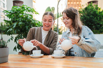 Two young women enjoying tea outdoors, smiling and bonding while pouring tea from white teapots at...