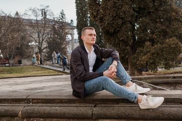 Fototapeta premium Young man sitting thoughtfully on a stone ledge in a park setting