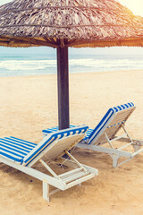 Beach lounge chairs with striped cushions under straw umbrella on sandy shoreline