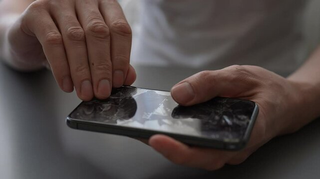 Skilled repairman removing shattered tempered glass screen protector from cellphone, revealing intact phone screen underneath in restore workshop, close-up. Shooting in slow motion.