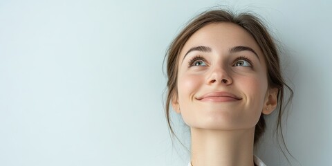 Naklejka premium Cheerful young woman with long brown hair smiling and looking upwards against a soft light blue background showcasing positivity and optimism