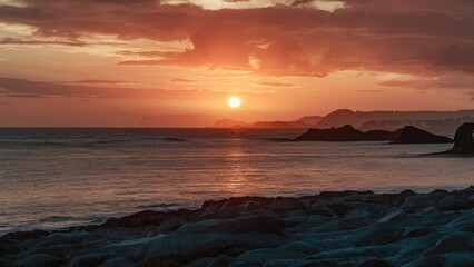Obraz premium Serene beach sunrise with orange and pink hues illuminating the sky as the sun rises above gentle waves and coastal silhouettes in the distance.