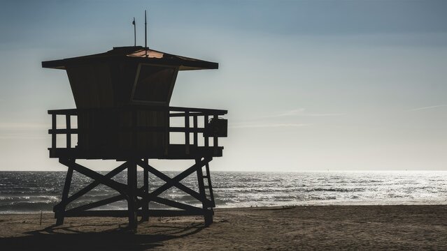 Silhouette of a lifeguard tower on a sandy beach with a shimmering sea in the background under a clear sky during golden hour lighting. - Powered by Adobe