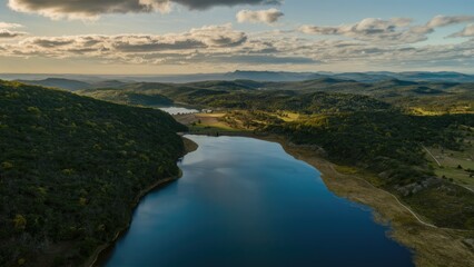 Aerial view of serene lake surrounded by lush green hills and scattered clouds under a golden sunset light showcasing natural beauty.
