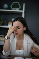 A businesswoman sits at her desk, visibly stressed and frustrated, rubbing her forehead as she holds her glasses in one hand. The office setting includes a laptop, a cup of coffee, and some paperwork