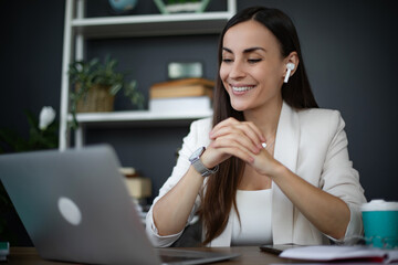 A smiling young woman with earphones, dressed in a white blazer and top, is sitting at a desk in an office, using a laptop and looking relaxed while engaged in a virtual meeting or conversation.