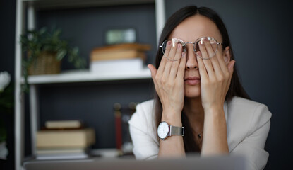 A woman in a modern office setting, wearing glasses and a white blazer, is visibly stressed or...