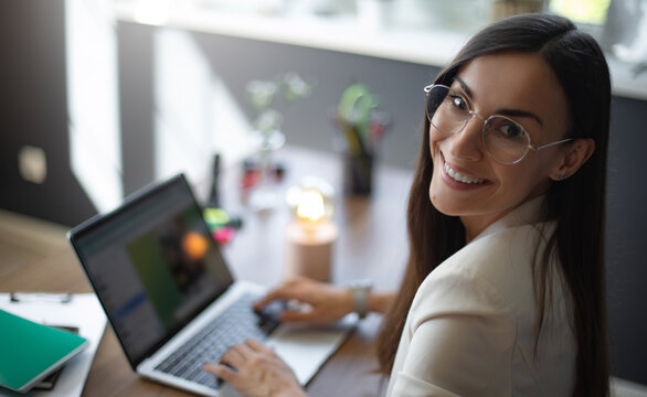 A smiling young woman in a white blazer sits at a desk, working on her laptop in a modern office space. She wears glasses and is surrounded by office supplies, plants, and a warm light on the desk.