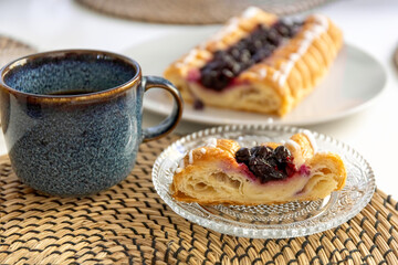 A slice of pie with berry filling on a glass plate and a mug of coffee on a dining table. Photo
