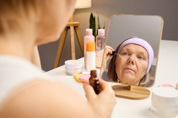 An elderly woman sits at a table in her home, carefully applying mascara while looking into a...