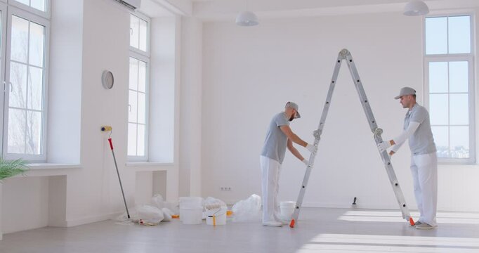 Two professional contractors in work uniforms carrying ladder across bright room while preparing for renovation. Male painters discussing design plans and organizing tasks for remodeling new house.