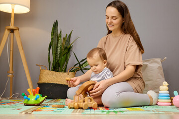 A mother and her baby sit on a colorful play mat in a cozy living room. They are focused on playing...
