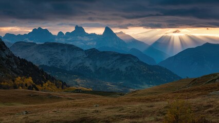 Obraz premium Majestic mountain peaks under a dramatic moody sky with sun rays piercing through clouds during autumn foliage creating a breathtaking landscape view