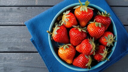 Vibrantly ripe strawberries arranged in a turquoise bowl on a blue napkin placed on a rustic wooden surface showcasing rich red colors.
