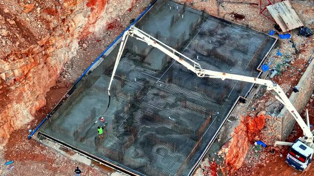 The image shows a foundation being built, with concrete poured via a pump and workers on-site. It reflects the early stages of construction progress in the project.
