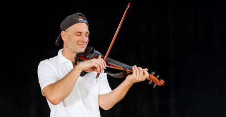 40 years old man in white shirt and black cap standing on the black background having joy while playing violin, musical instrument in scene. Male violinist, copy space
