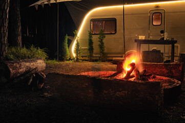Glowing campfire surrounded by logs in front of a warmly lit camper, creating a cozy night camping...