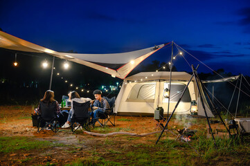 Outdoor camping tent in the forest park, party dinner with friends under tarp or flysheet and warm lighting at night near natural under a twilight sky. Overview of travel camping with friends.