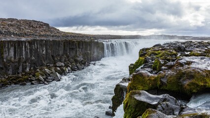 Scenic view of Dettifoss waterfall in Iceland with rugged cliffs and mossy rocks under a cloudy sky.