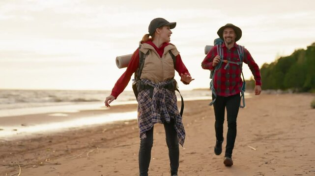 Active pastime in weekend, happy man and woman traveling to seacoast, running. Slow motion shot with joyful backpackers enjoying joint time, laughing hikers running on beach, good mood and positivity