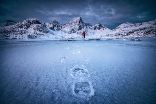 Scandinavian winter Landscape of Snowy Mountain peaks and sea coast, frozen lake water with footsteps in snow and photographer person with camera