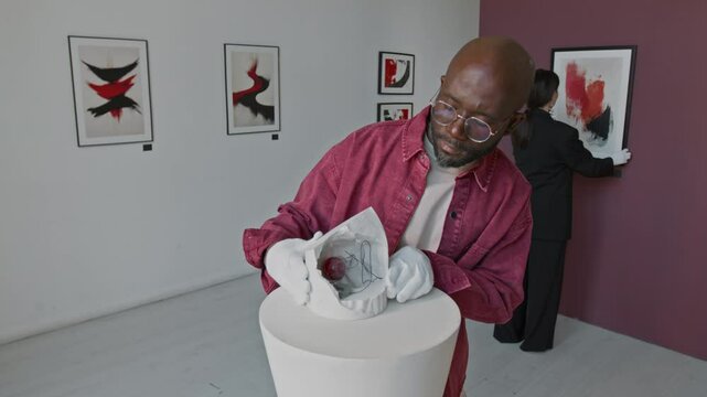 African American man carefully placing abstract instalation on pedesnal, while his female colleague adjusing painting on wall in minimalist art gallery