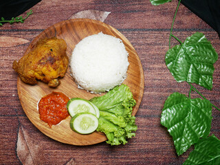 Top view traditional Indonesian food consists of white rice, fried chicken, spicy red Sambal, fresh cucumber slices, and lettuce leaves on a round wooden plate. Decorated with green plastic leaves.