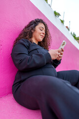 Pregnant woman sitting relaxed, scrolling smartphone against bright pink backdrop, capturing serene...