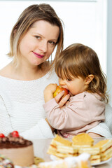Cute toddler eating apple, prefer fresh fruits against sweets sitting behind table with sweet food