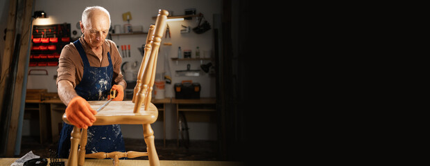 Senior carpenter measure old wooden chair with measurement tape in dark carpentry workshop. Furniture restoration, DIY and hobby concept