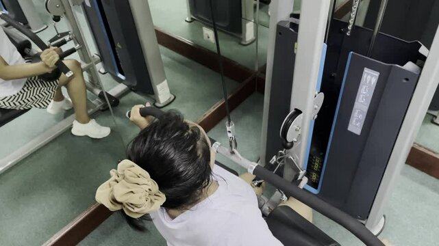 A woman is using the lat pulldown machine at the gym. She is pulling the bar down with both hands. She wears a white t-shirt and a patterned skirt. Her hair is black, and she has a tan scrunchie on.