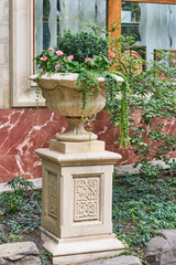 Classical stone urn on carved pedestal with ornate details. Pink flowers and trailing greenery