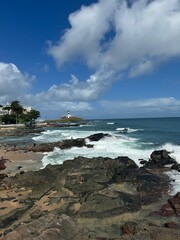 Beautiful coastal view featuring the Barra Lighthouse in Salvador, Brazil. Waves crashing against the rocky shoreline under a bright blue sky filled with fluffy clouds. A serene tropical scene
