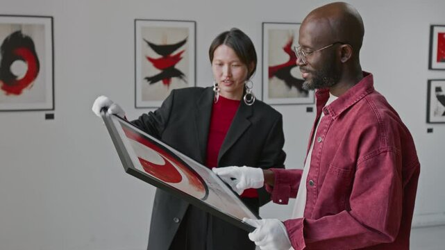 Asian woman and Black man holding framed abstract painting together and discussing it, while setting up exhibition in minimalist art gallery