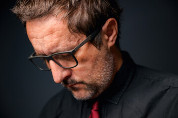 Fototapeta premium Portrait of serious concerned businessman entrepreneur wearing eyeglasses and black shirt in dark office interior, closeup low key