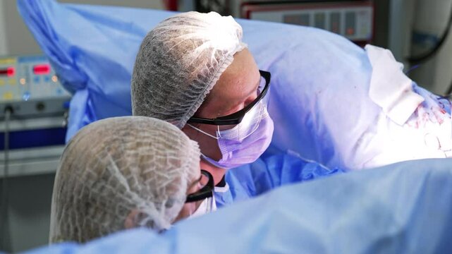 Shot over the head of two female surgeons wearing caps, protective glasses and masks. Doctors sit at the legs of the patient carrying out gynecology operation.