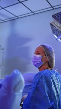 Attentive female surgeon performs operation looking at monitor. Flashes from the screen can be seen on doctor's face. Professional uses tool device at gynecologic surgery. Vertical video.