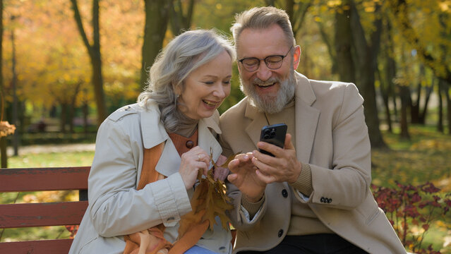 Caucasian retired couple family in love mature woman man sit on bench autumn park using mobile phone gadget together smiling wife husband spouses reading news old age and smartphone in city outdoors