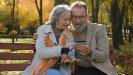 Married Caucasian couple happy family middle-aged mature woman man remote paying online order with mobile phone credit card retired husband wife using smartphone bank app shopping in autumn park city