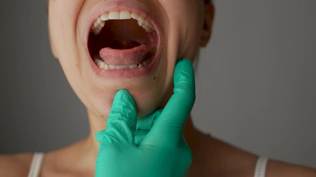 Doctor's hand in a green glove opens the female patient's mouth by examining the inflamed red throat on dark grey background. Examination at the therapist. Flu, cold, tonsillitis, sars virus