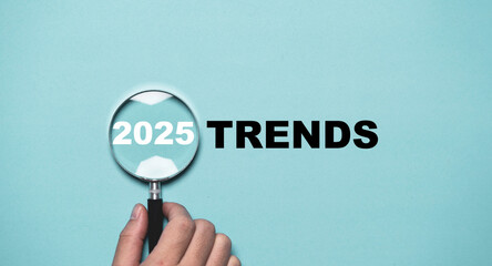 Hand holding magnifier glass with 2025 trends on blue background for searching and presents the evaluation methods, popular topics in marketing monitor and business planing in new year concept.
