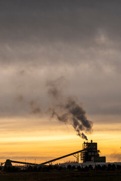 An industrial factory belches thick smoke into the sky at sunset, illustrating concerns about pollution and environmental impact in an urban landscape.