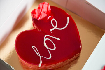 Close-up of a glazed red heart-shaped cake with the inscription "Love" is placed in a box on a light background. The theme is Valentine's Day.
