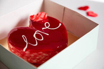 A glazed red heart-shaped cake with the inscription "Love" is placed in a box on a light background. The theme is Valentine's Day.
